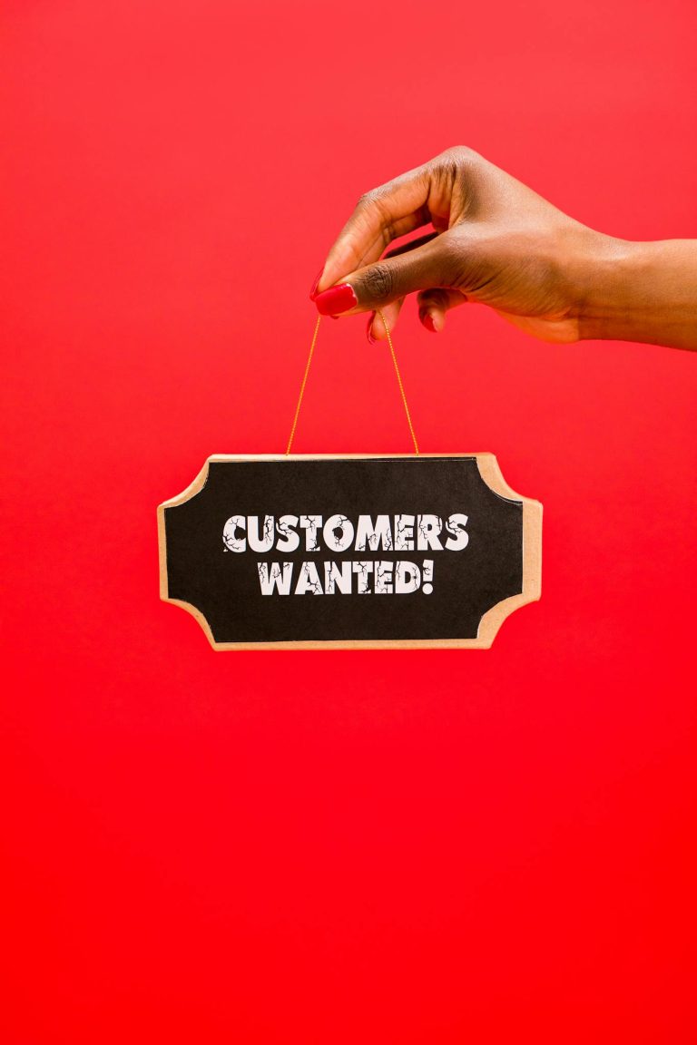 Close-up of a hand holding a 'Customers Wanted' sign against a bright red background.